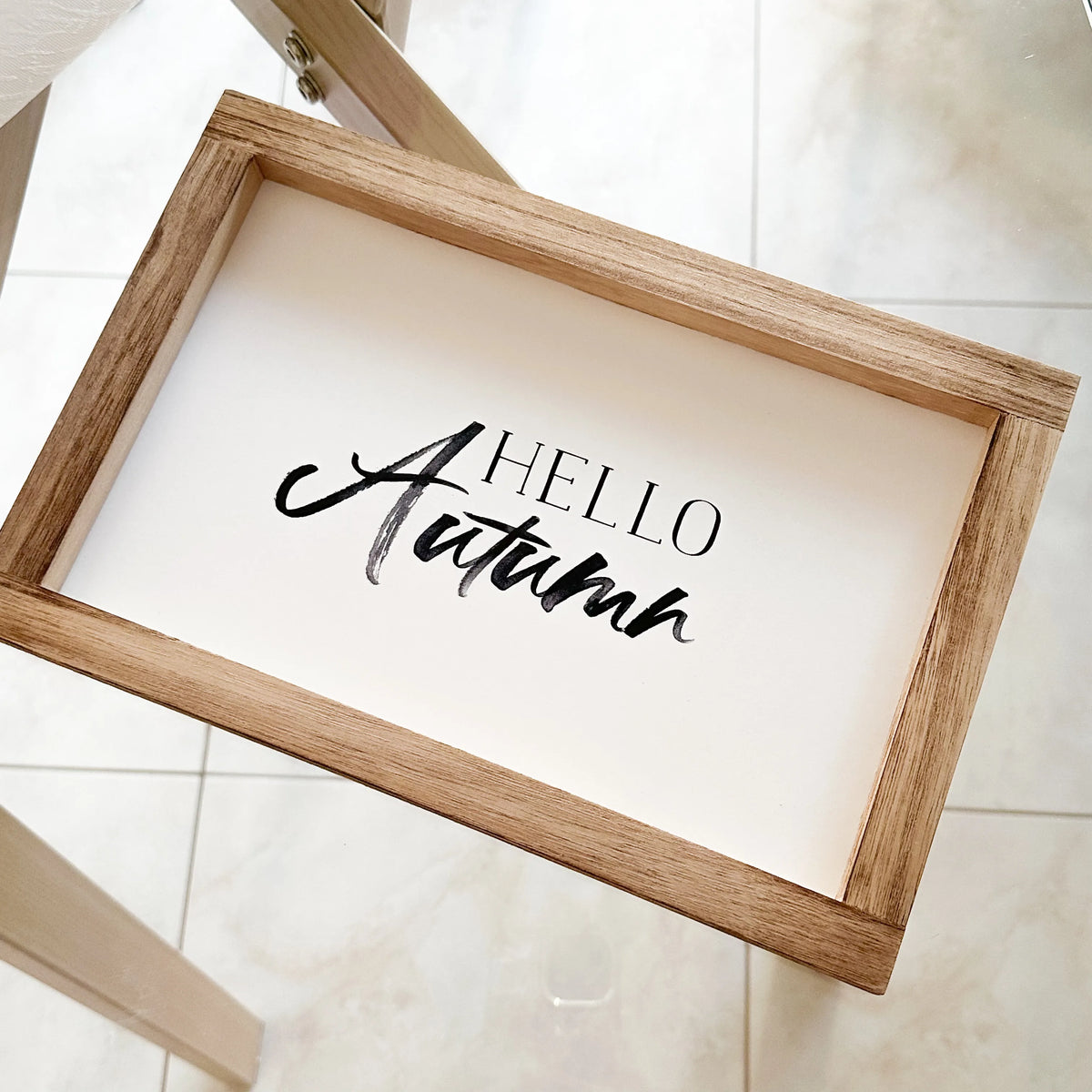 Quadretto Hello Autumn con cornice in legno naturale chiaro e fondo in legno bianco con scritta Hello Autumn in stile moderno, vista dall'alto su pavimento in gres chiaro