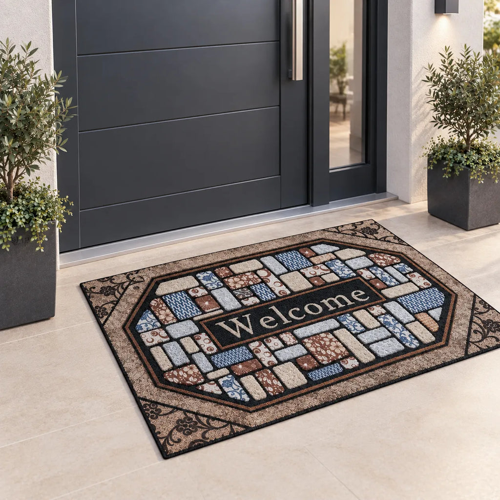 Zerbino Essential posato davanti a una porta d'ingresso grigia, con decoro a rilievo che imita lastre di pietra colorate in bronzo, bianco, marrone e celeste e scritta Welcome su fondo nero