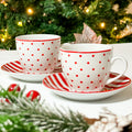 Set di tazze natalizie bianche a pois rossi con piattini a righe rosse e bianche, davanti a un albero di Natale